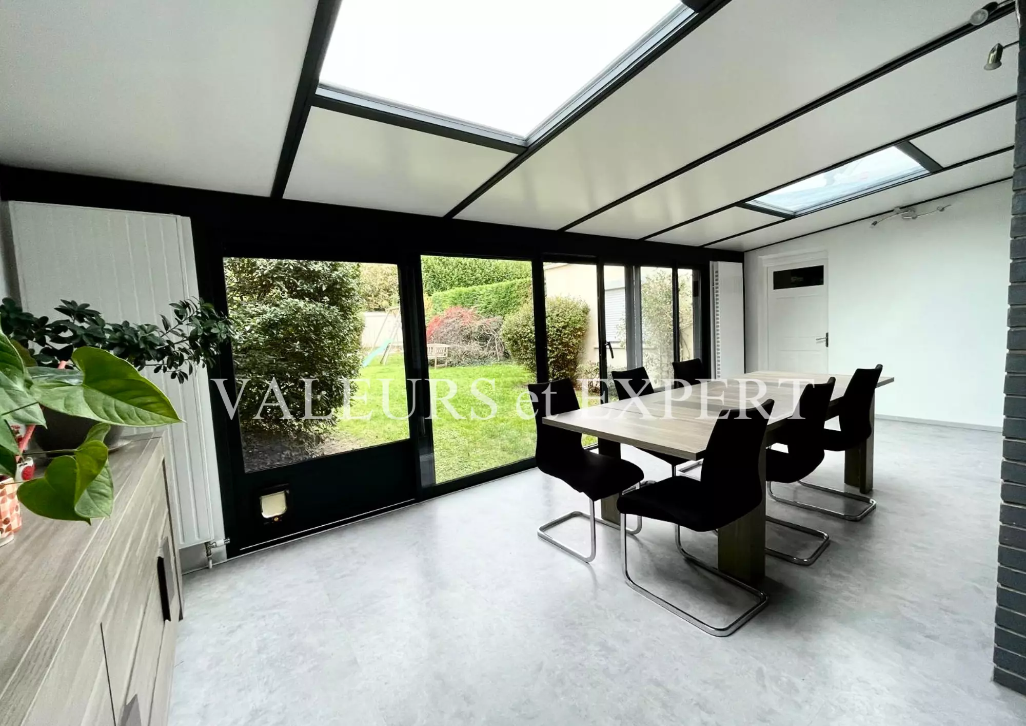 Salle à manger lumineuse avec véranda et grandes baies vitrées donnant sur le jardin.